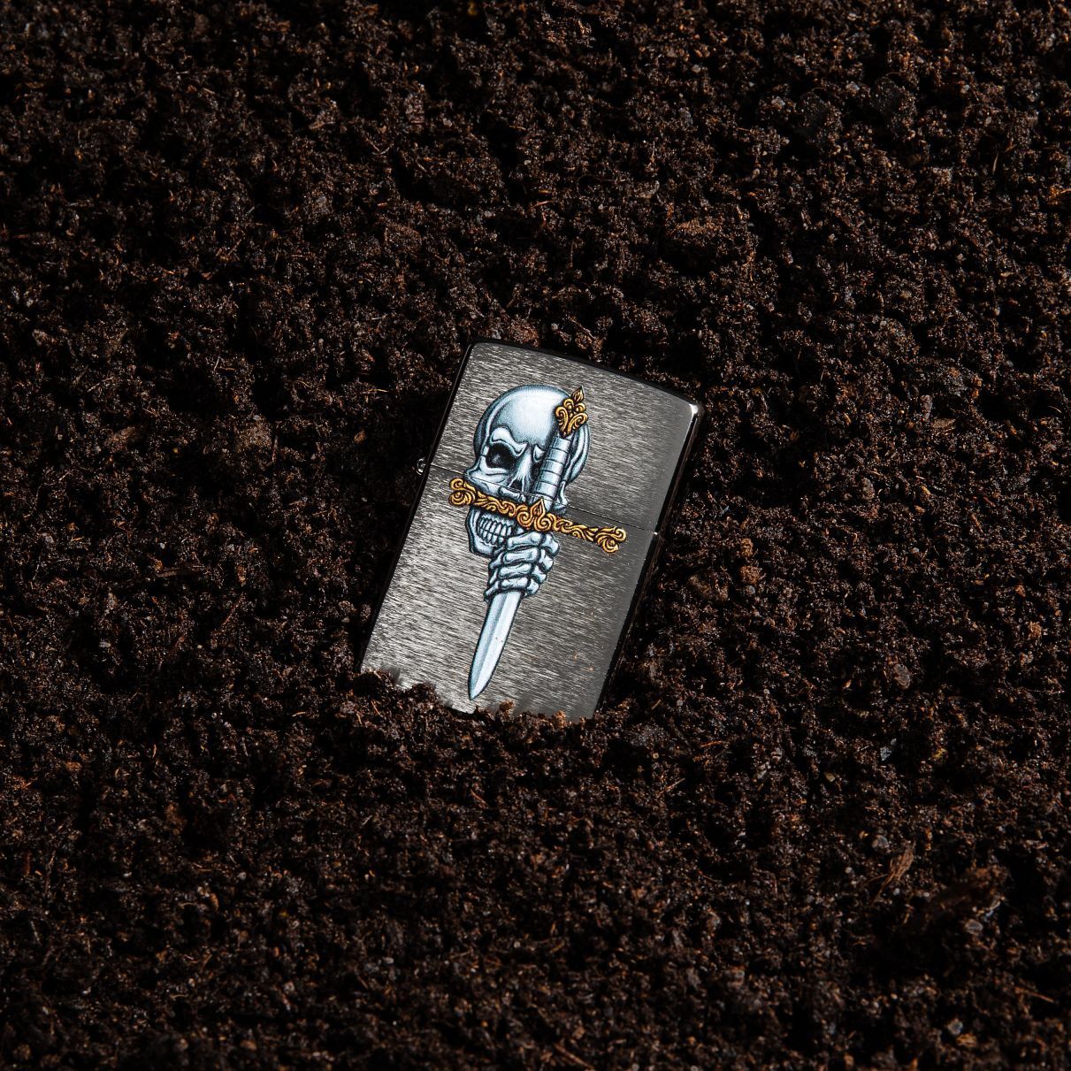 Lifestyle image of Sword Skull Design Brushed Chrome Windproof Lighter laying in dirt.